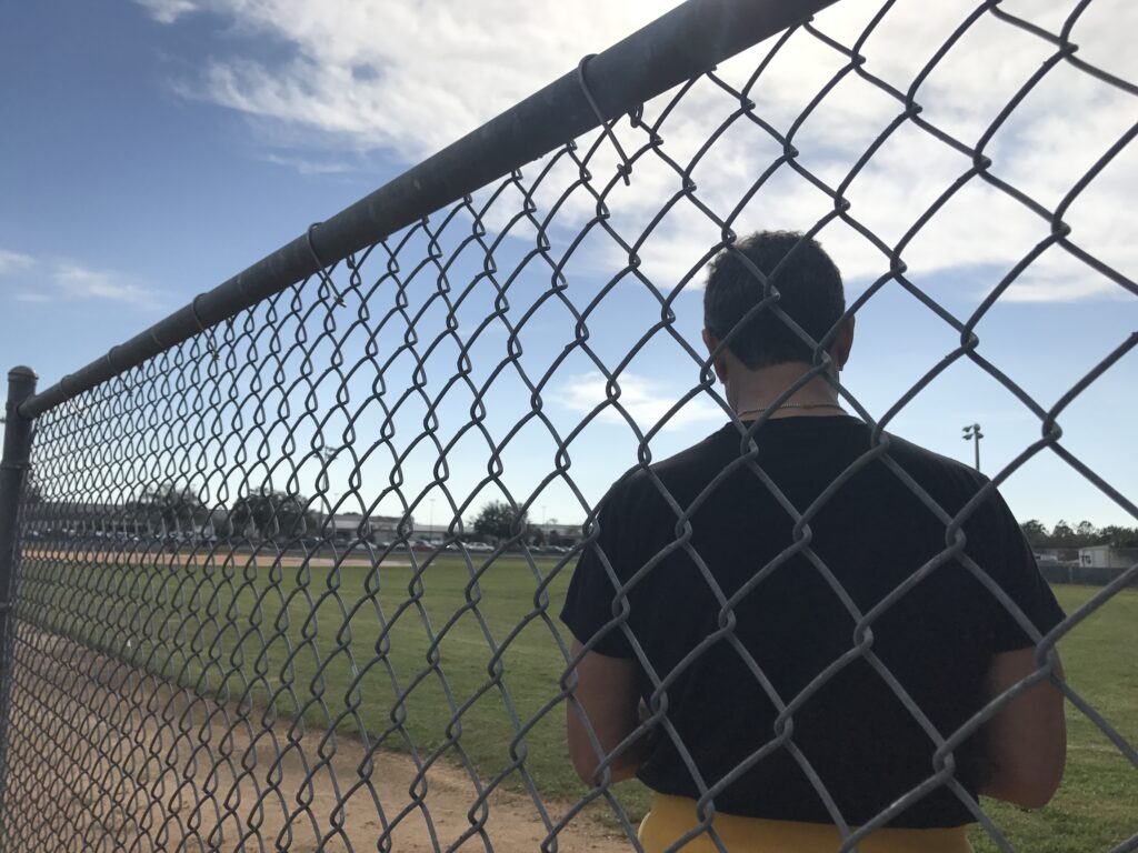 man, with back to camera, sitting next to chain link fence